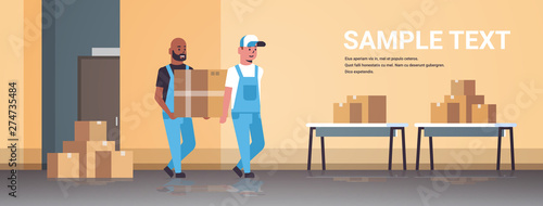 two couriers in uniform carrying cardboard box package mail express delivery service concept mix race industrial factory workers working in stockroom of storehouse flat full length horizontal
