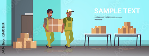 two couriers in uniform carrying cardboard box package mail express delivery service concept african american industrial factory workers working in stockroom of storehouse flat full length horizontal