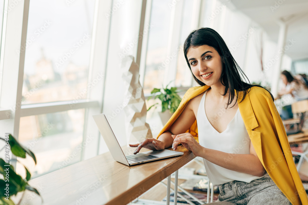 Beautiful attractive young georgian woman sit at table in coworking. Typing on laptop keyboard. Posing on camera and smile. Alone in white room. Beautiful attractive young georgian woman sit at table in coworking. Typing on laptop keyboard. Posing on camera and smile. Alone in white room.
