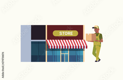 postman in uniform carrying cardboard parcel box african american courier express delivery service concept modern store building exterior flat full length horizontal