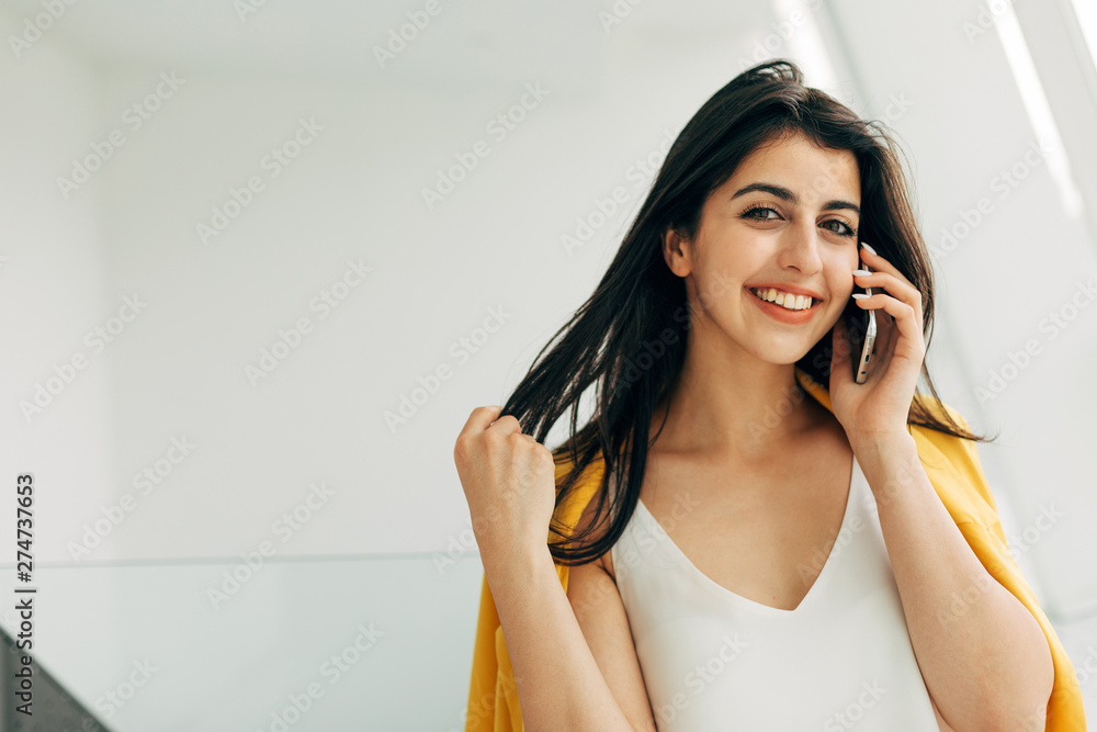 Fototapeta premium Picture of happy attractive young woman look straight on camera and smile. Touch hair and talk on phone. Businesswoman in deal. Stand at white wall or window.