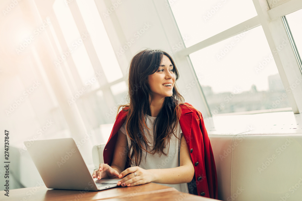 Cheerful attractive young buisnesswoman in white room. Remote work on project. Look back and smile. Sit in coworking place at window. Work alone. Wear stylish red jacket. Cheerful attractive young buisnesswoman in white room. Remote work on project. Look back and smile. Sit in coworking place at window. Work alone. Wear stylish red jacket.