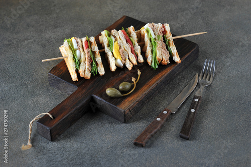 Tuna sandwich on a wooden spit. Sandwich served on a wooden board. Near cutlery and capers. Dark background. Close-up.
