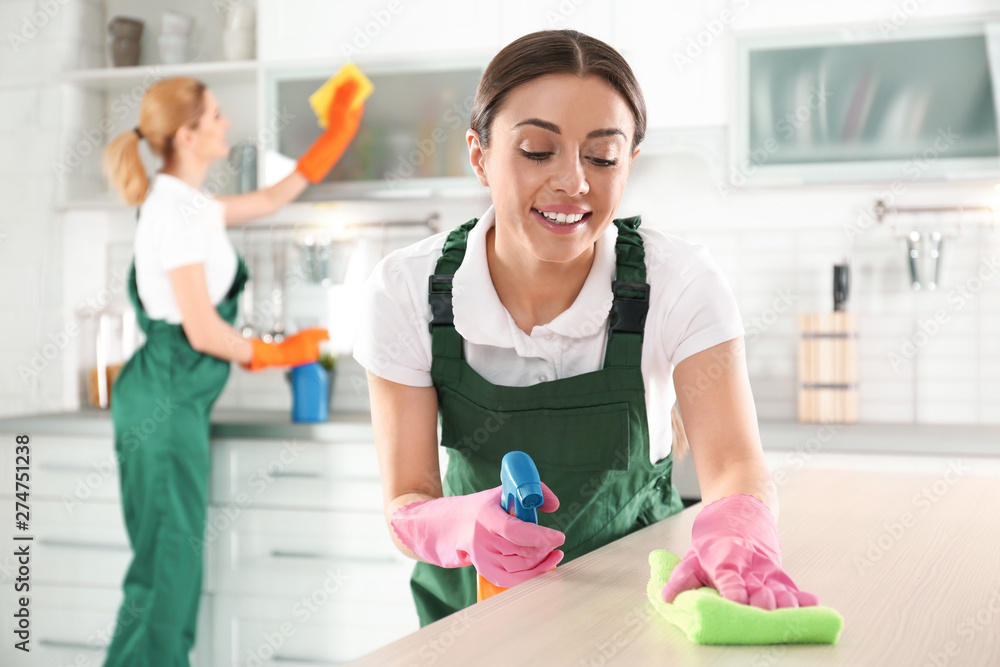 Woman using rag and sprayer for cleaning table with colleague in kitchen Woman using rag and sprayer for cleaning table with colleague in kitchen
