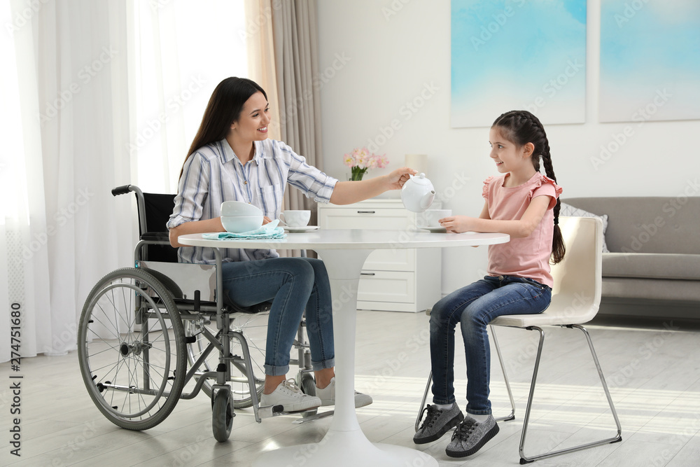 Mother in wheelchair and her daughter drinking tea at table indoors Mother in wheelchair and her daughter drinking tea at table indoors