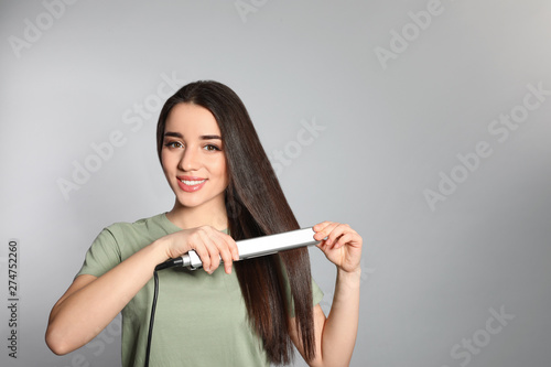 Happy woman using hair iron on grey background. Space for text