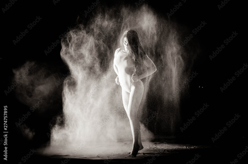 Obraz premium The girl with the flour on the body stretches the arms up with thrown flour on black background black and white image