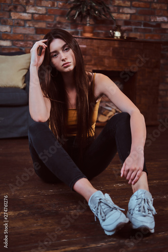 Attractive bored teenager is sitting on the floor in the room. She has long hair and white sneakers.