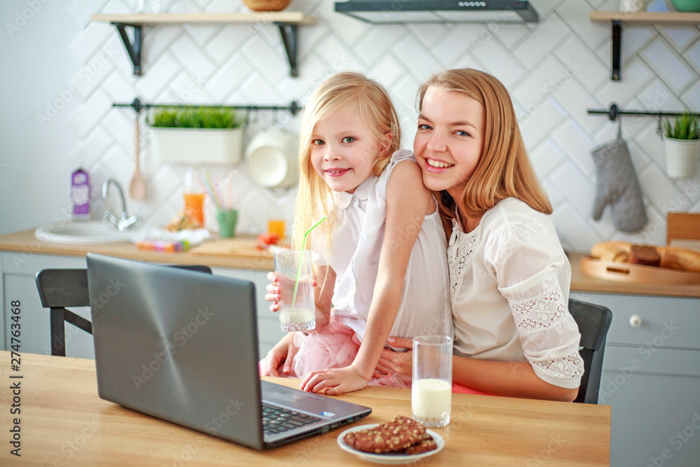 Mother with baby daughter sitting at table with groceries in kitchen with laptop