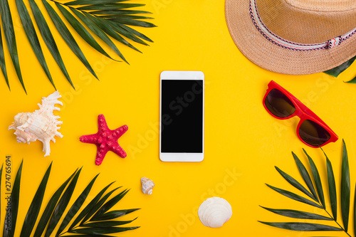 Hat, tropical palm leaves, sunglasses, sea shells and smartphone on yellow background.