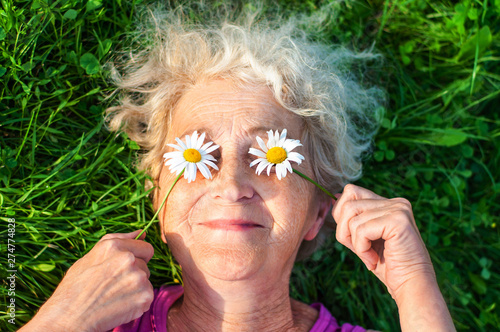 Mature woman after 60 is lying on the lawn holding chamomile eyes. Care for women's wrinkled skin. Vision and eye care. Women Health