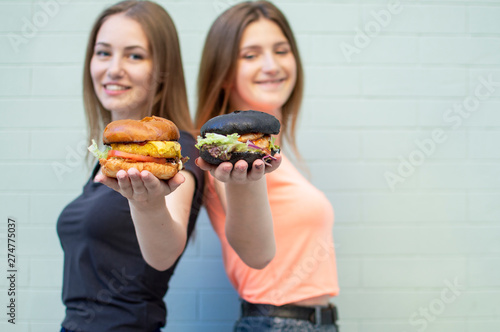 young beautiful girls-students stand near the blue wall, hold burgers and smile, they are happy and show cheeseburgers on camera