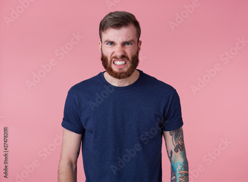 Portrait of young handsome angry red bearded man in red t-shirt, stands over pink background, looks at the camera with disgust and bares his teeth.