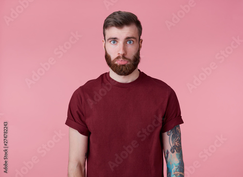 Quadro em tela Portrait of sad young handsome red bearded tattooed man in red t-shirt, offended looking at the camera with a lowered lip, stands over pink background