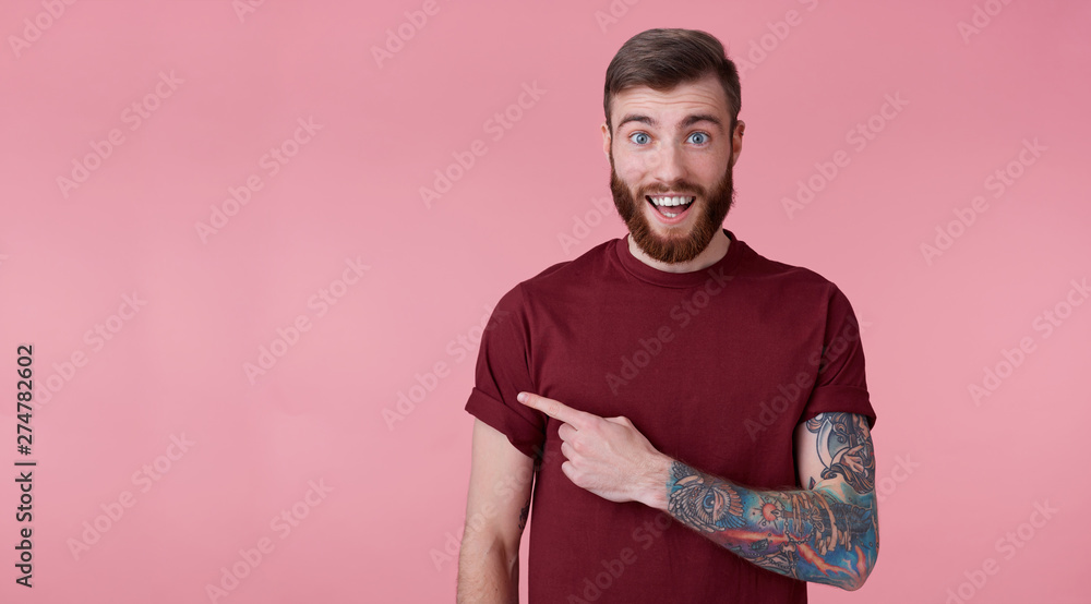 Young handsome happy amazed red bearded man in blank t-shirt, looks surprised, stands over pink background broadly smiling, wants to draw you attention and points to copy space on the left side. Young handsome happy amazed red bearded man in blank t-shirt, looks surprised, stands over pink background broadly smiling, wants to draw you attention and points to copy space on the left side.
