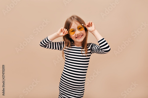 Funny happy 6 years old girl in stripped dress wearing round orange glasses looking away with charming smile over beige background 