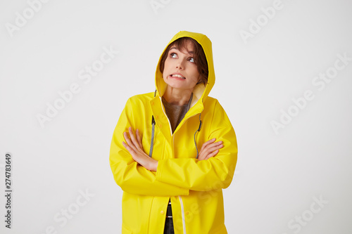 Portrait of young disgusted cute short haired girl wears in yellow rain coat, looks up with unhappy expressions, hiding under a rain hood, stands over white wall with crossed arms. photo