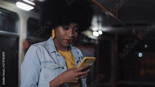 Attractive s,iling young african american woman using smartphone at public transport. Night time. City lights background. photo