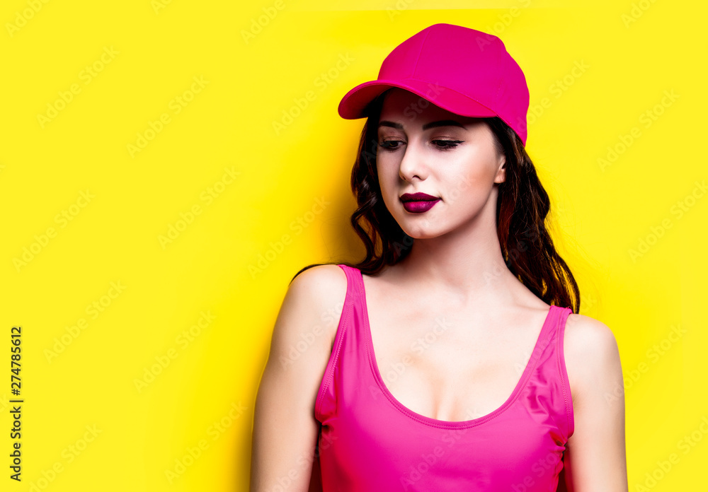 Elegant brunette woman in pink swimsuit and fashion cap. Sexy lady in pink beachwear, sunglasses enjoying sun on yellow summer background Elegant brunette woman in pink swimsuit and fashion cap. Sexy lady in pink beachwear, sunglasses enjoying sun on yellow summer background