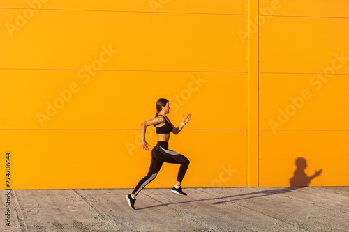 Side view of young attractive sporty woman wearing black sporwear practicing sport exercises in morning on street, start to run with speed, running silhouette, orange wall background, outdoor