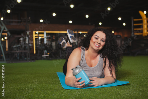 Cheerful plus size woman relaxing at the gym after working out, lying on yoga mat, copy space. Happy beautiful overweight sportswoman resting at sport studio