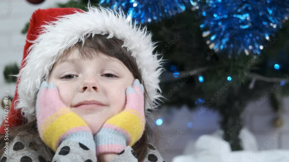 Happy child near the Christmas tree. Portrait of a cute little girl in a santa claus hat against the background of a christmas tree. Waiting for Christmas. Happy child near the Christmas tree. Portrait of a cute little girl in a santa claus hat against the background of a christmas tree. Waiting for Christmas.