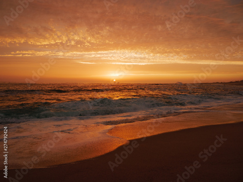 Waves crash on sandy beach at sunset with vivid orange sky