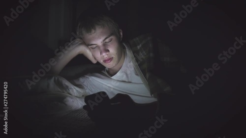 Bored teenager lying on his bed in the dark while scrolling through his phone. The light from the phone is illuminating his face.