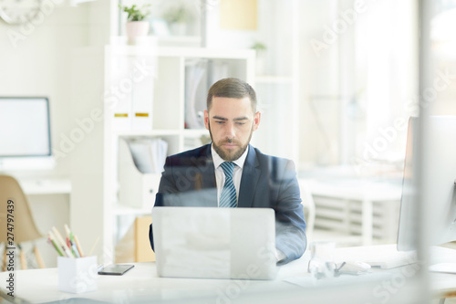 Serious busy young businessman with beard sitting at desk in office and working with laptop