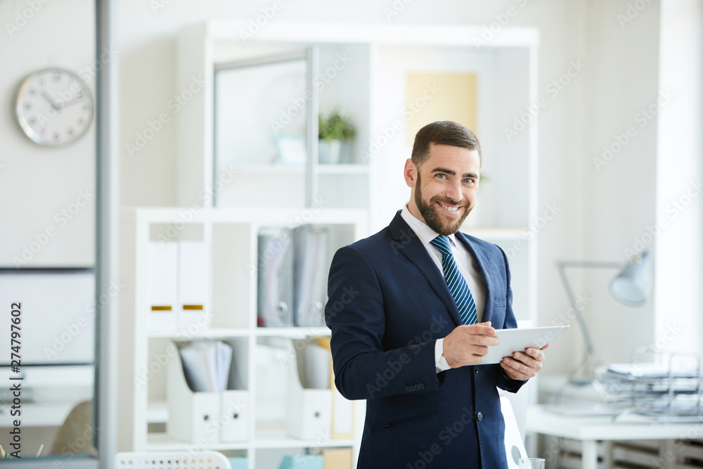 Portrait of cheerful confident handsome young manager in dark suit standing in office and using digital tablet