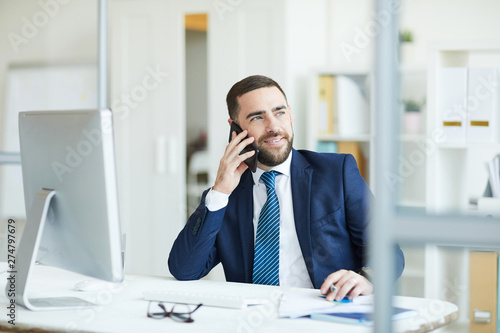 Positive confident handsome young project manager with beard sitting at table and discussing plans with colleague on mobile phone