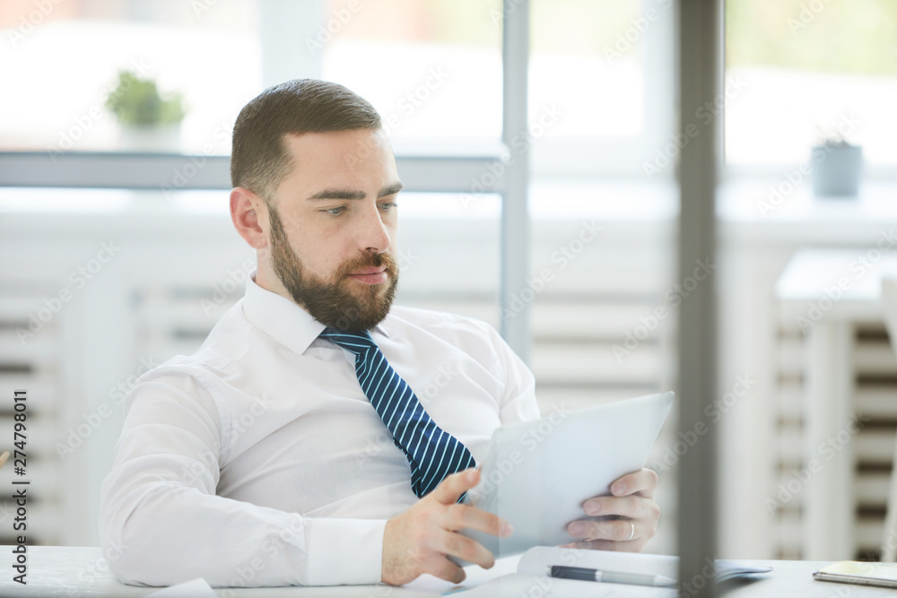 Serious busy young bearded manager sitting in relaxed pose at table in office and using tablet while checking data Serious busy young bearded manager sitting in relaxed pose at table in office and using tablet while checking data