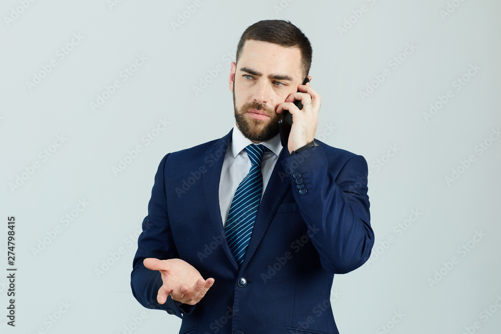 Serious displeased young bearded businessman in formal suit standing against white wall and gesturing hand while talking on mobile phone Serious displeased young bearded businessman in formal suit standing against white wall and gesturing hand while talking on mobile phone