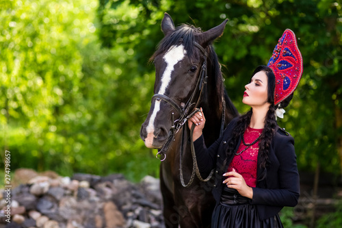 Lovely Fashion Model in Russian Style Kokoshnik Holding Thoroughbred Horse Closely.Posing Against Nature Background.