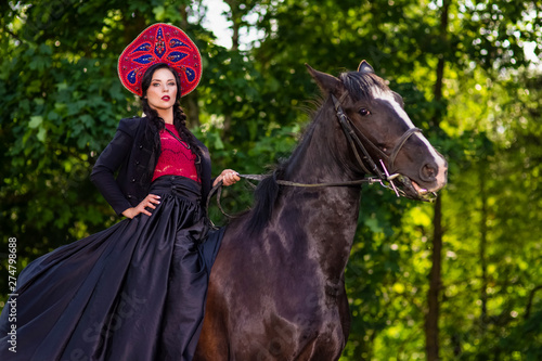 Gorgeous Fashion Model in Russian Style Kokoshnik Straddling On the Thoroughbred Horse. Posing Against Nature Background