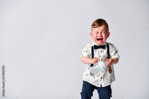 Crying baby. Portrait of a kid in full growth in the studio on a white background. Dressed in a suit of a gentleman - white shirt and pants with straps. © FAB.1