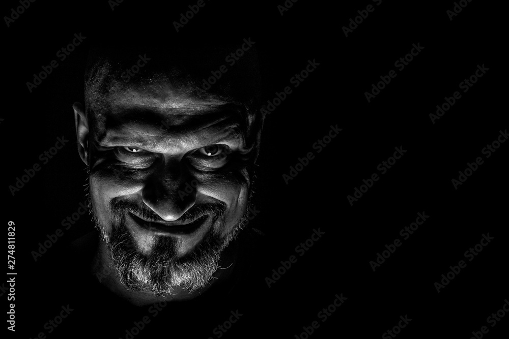 Face with a bearded man grimace against a dark background with sharp shadows. Comedic, fabulous villain or negative character conception with copy space. Face with a bearded man grimace against a dark background with sharp shadows. Comedic, fabulous villain or negative character conception with copy space.