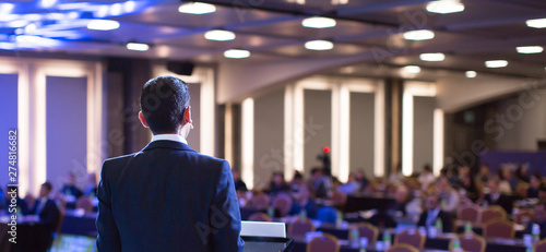 Speaker giving a talk at a corporate business conference. Audience in hall with presenter in front of presentation screen. Corporate executive giving speech during business and entrepreneur seminar.
