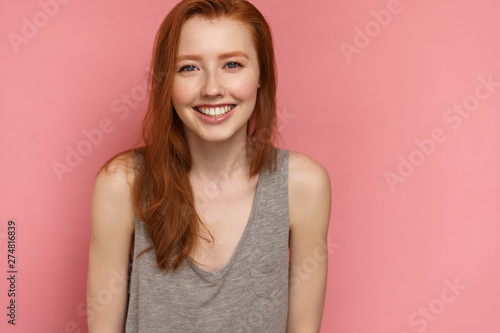 Red-haired young woman smiles at the camera posing over pink background, close-up