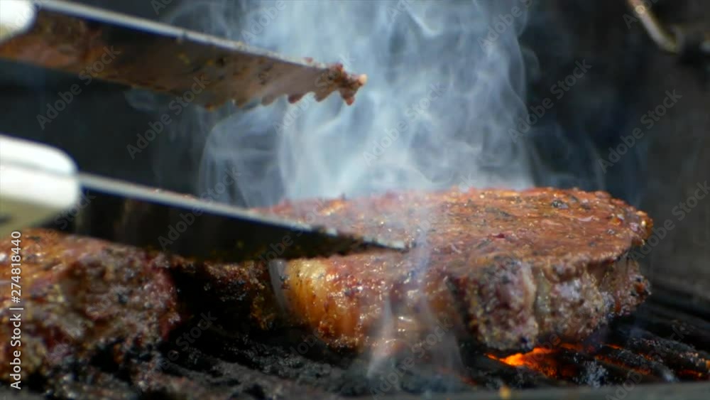 A pair of meat tongs turn a nearly cooked juicy rib eye steak on a grill as orange flames shoot up from below. A pair of meat tongs turn a nearly cooked juicy rib eye steak on a grill as orange flames shoot up from below.