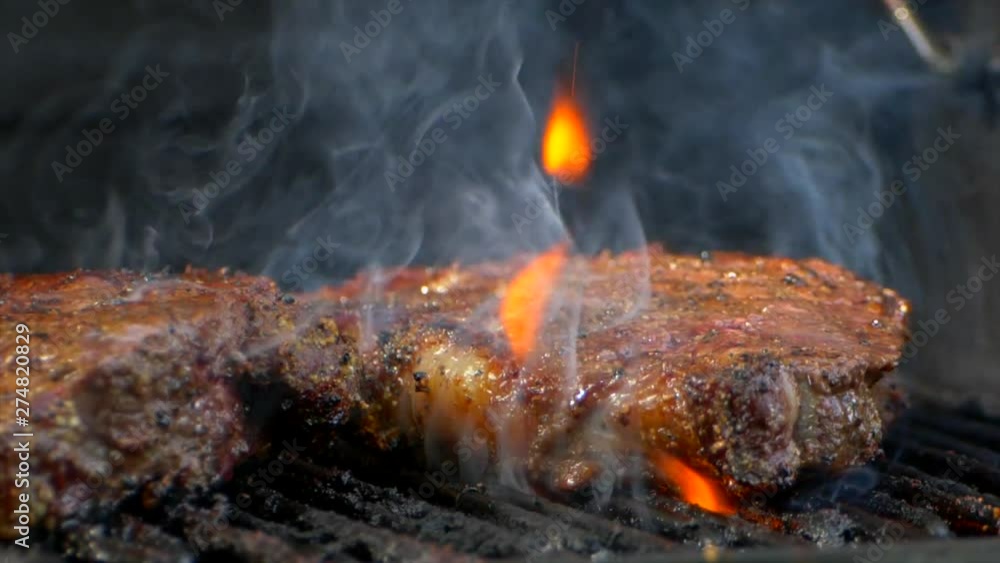 A pair of meat tongs slide a nearly cooked juicy rib eye steak on a grill and presses on it as flames shoot up. A pair of meat tongs slide a nearly cooked juicy rib eye steak on a grill and presses on it as flames shoot up.