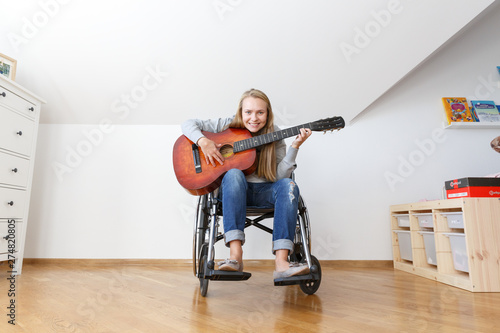 Disabled young woman in wheelchair playing the guitar. Wide angle, full body.