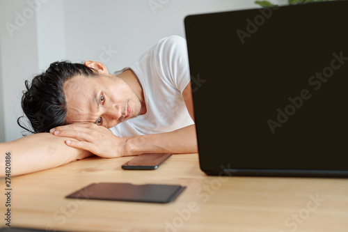 Bored young Asian man leaning on table when waiting for video rendering