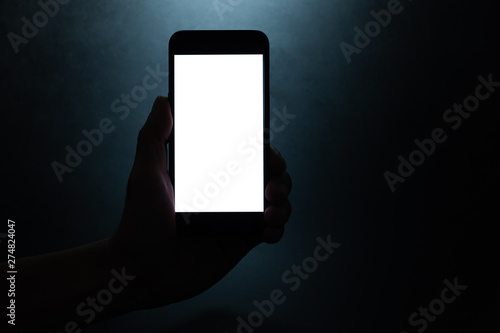 silhouette hand holding a mobile phone with blank wihte screen in the dark darkness against dark blue background