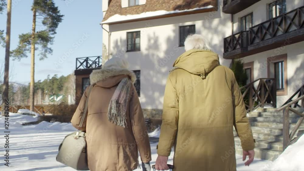 Tilting rear follow shot of mature couple, wearing warm parkas, arriving at countryside hotel on bright winter’s day, chatting and wheeling suitcase, then going up stony steps towards entrance Tilting rear follow shot of mature couple, wearing warm parkas, arriving at countryside hotel on bright winter’s day, chatting and wheeling suitcase, then going up stony steps towards entrance