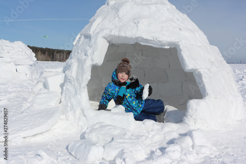 Happy cheerful boy with a piece of snow in his hands sitting an igloo in winter