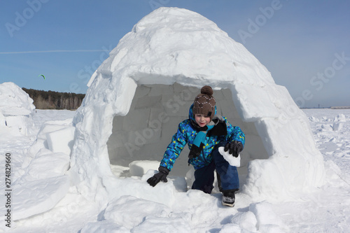 Boy with a piece of snow in his hands comes out an igloo in winter