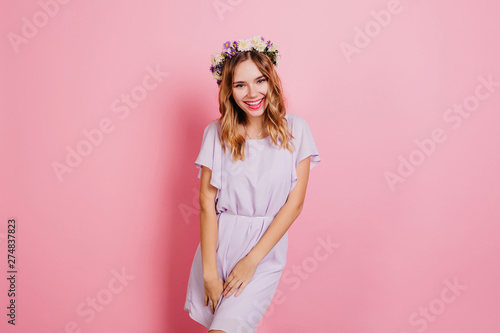 Lightly-tanned girl in trendy flower wreath laughing during photoshoot. Portrait of smiling blonde woman in summer dress isolated on pink background.