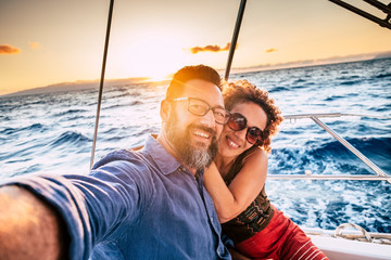 Happy and cheerful people enjoying the travel and trip on a sail boat with ocean and sunset sunlight in background - traveler lifestyle for adult man and woman smiling doing a selfie picture