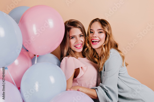 Laughing curly lady embracing her friend at party photoshoot. Portrait of girls with light-brown hair posing with colorful balloons on light background.